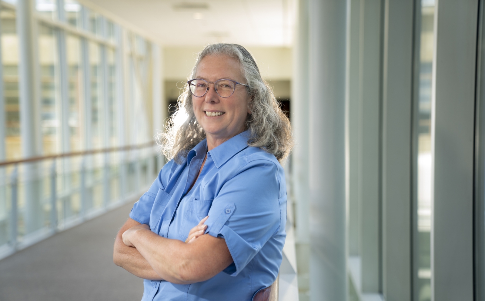Mary Ellen Zurko poses for a photo in a sunny, glass hallway. She's wearing a light blue shirt and her arms are crossed in front of her.