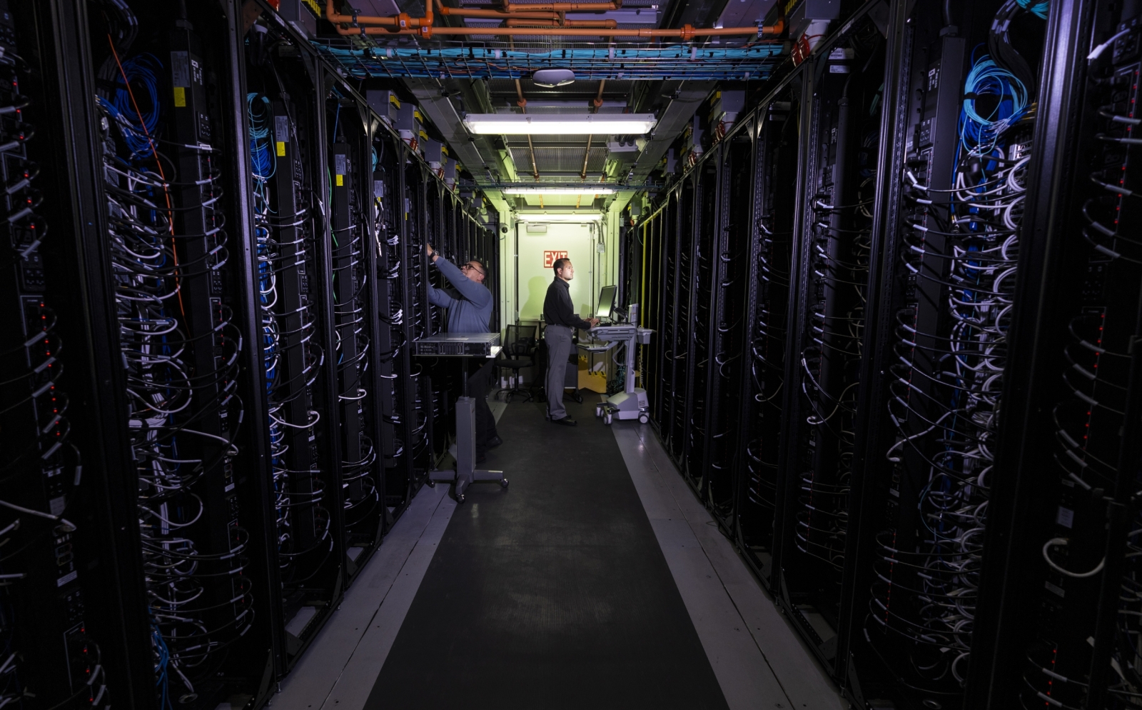 A photo of a hallway between two rows of data center servers. Two researchers are working on the servers.