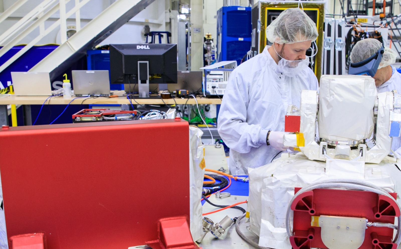 Two individuals in cleanroom attire examine an optical communications system payload. 