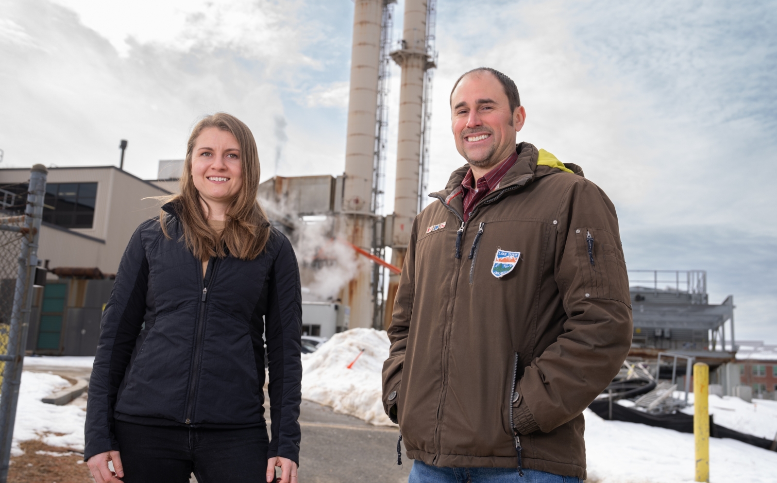 Jean Sack and Chris Lashway pose for a photo outside, with power-plant infrastructure in the background.