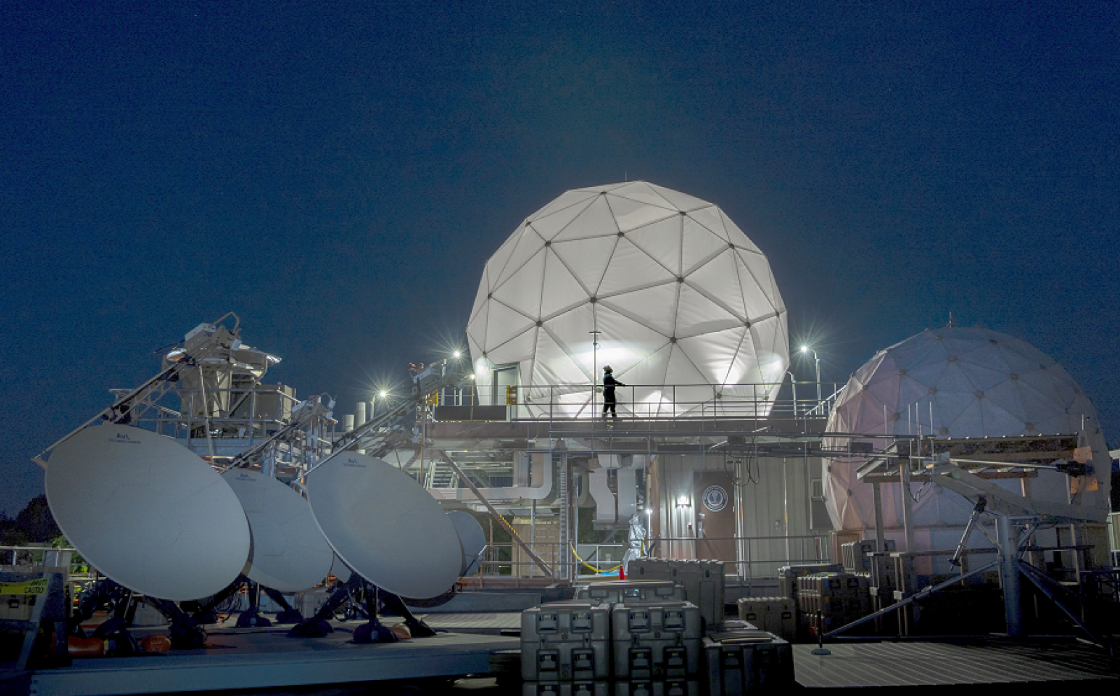 Four satellite dishes and two radomes, each covering satellite communications antennas, are shown on the roof of Lincoln Laboratory among other equipment.