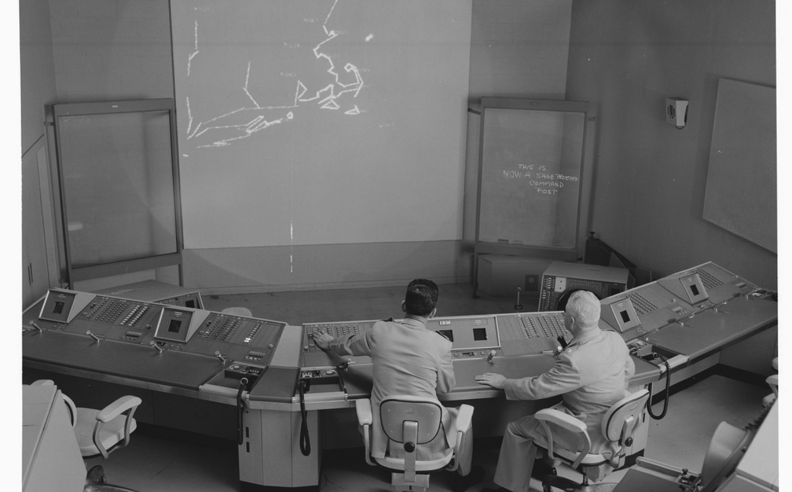 Two operators sit in front of a digital display showing an air defense network.