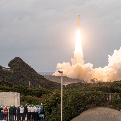 Spectators on a platform watch as the Quasi-Zenith Satellite System-Hosted Payload lifts off in the distance over a forest. 