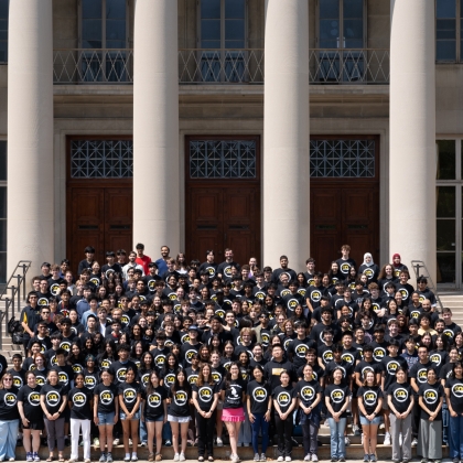 BWSI students wear matching black shirts with the BWSI logo and stand on steps outside of an MIT building with columns in the background. 