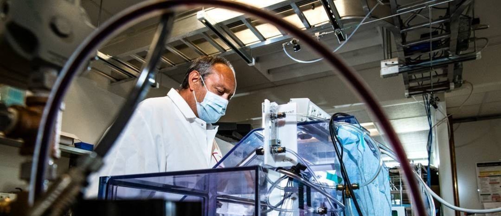 A man stands in a lab wearing a mask, in front of a big box setup where he is testing masks for air filtration. 