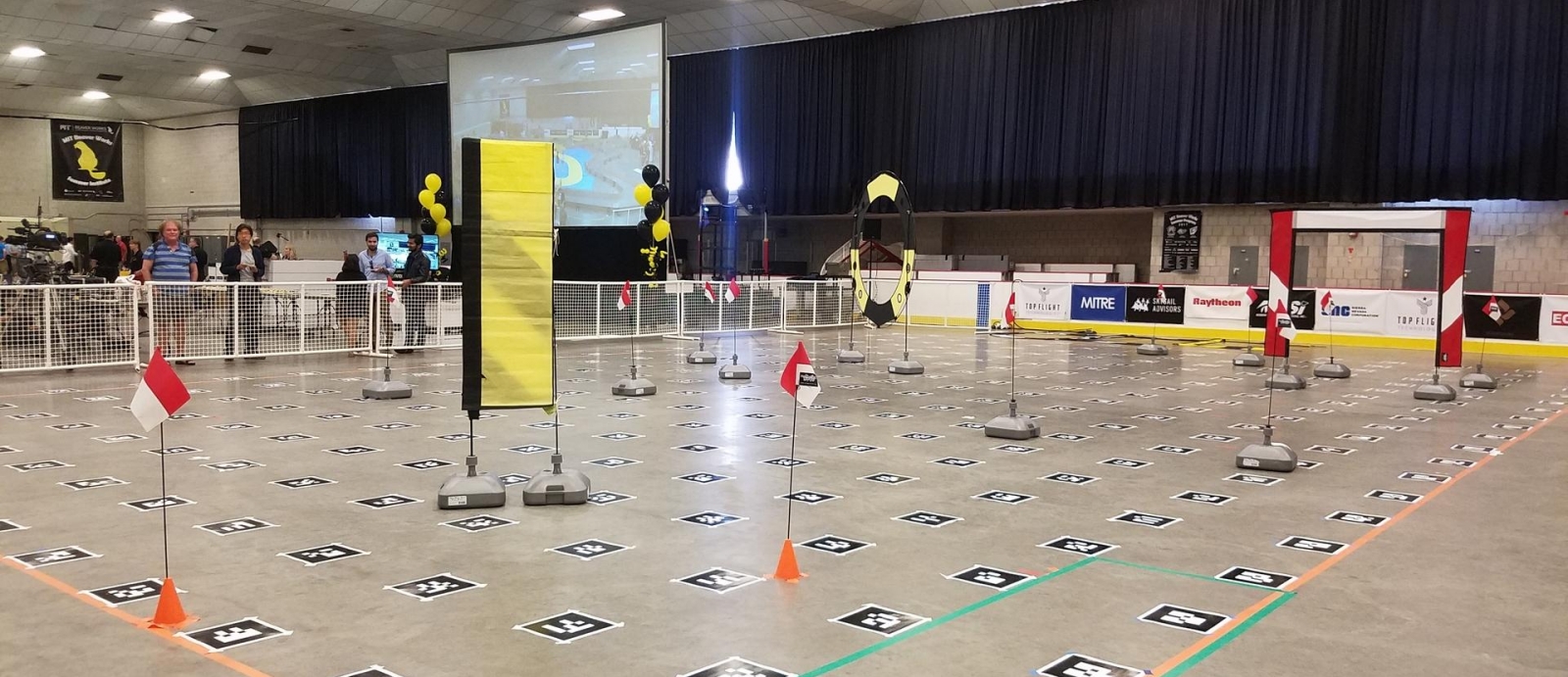 The race course for the Obstacles Trial. The three obstacles (from left) were a pillar, a hoop, and a gate. The flags indicate the required path that the drone must fly through while the black squares are Augmented Reality Codes for drones to detect.