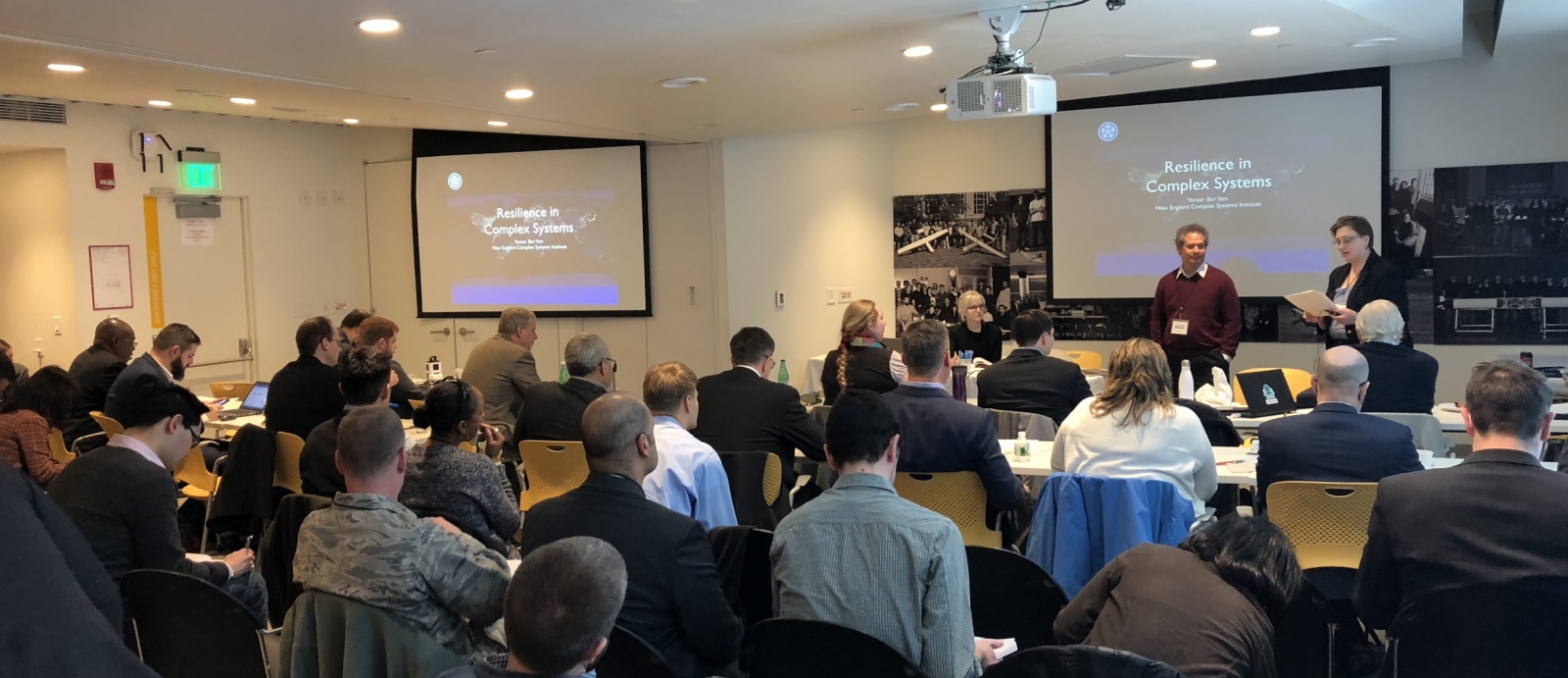 About thirty people sit in a classroom setting, with chairs facing a panel at front and projectors with a slide reading "Resilience in Complex Systems"