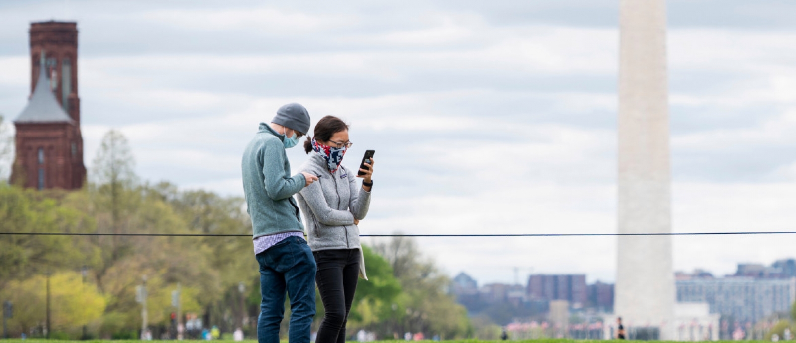 A photo of two people standing together wearing face masks, with the washington monument in the background. 