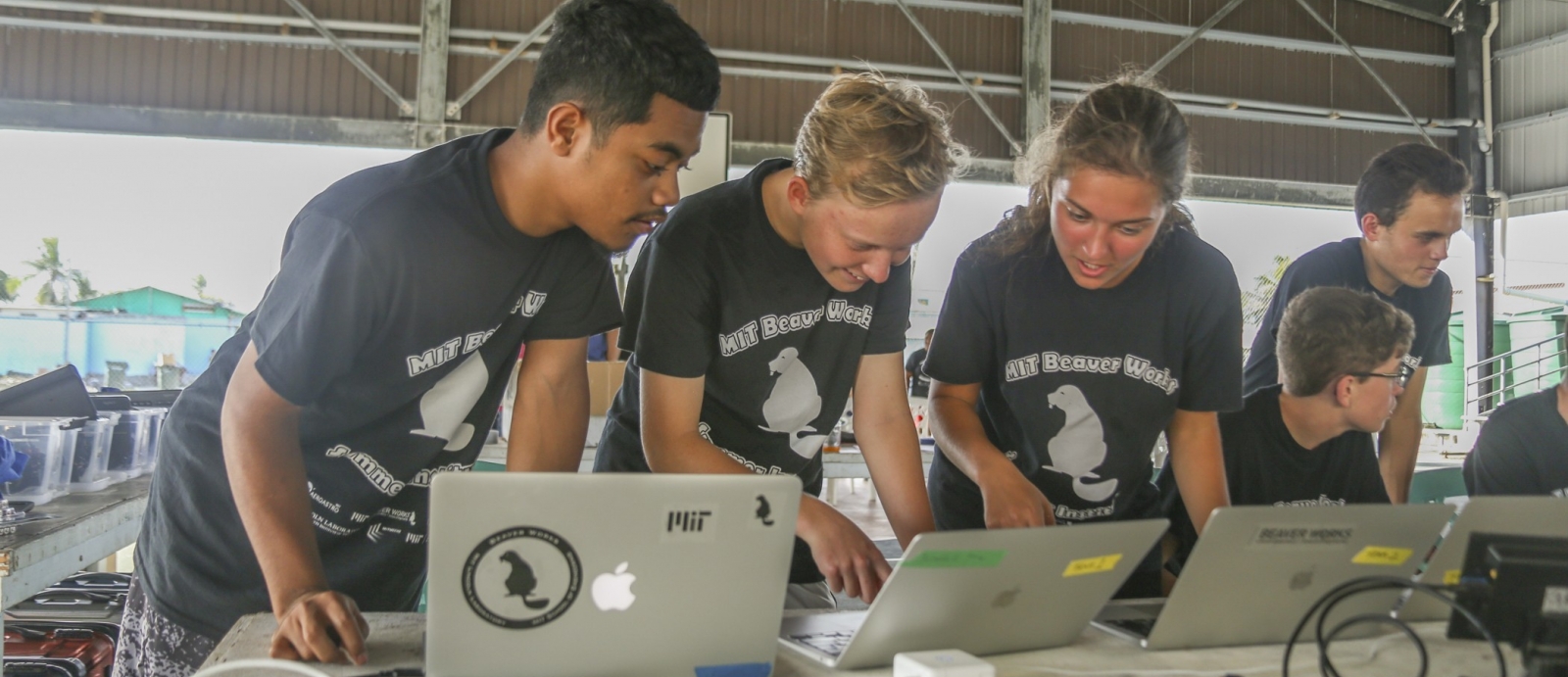 Three students stand together at a table, looking down each at a laptop, discussing. Two other students are in the background. The building they are in looks like an aircraft hanger.