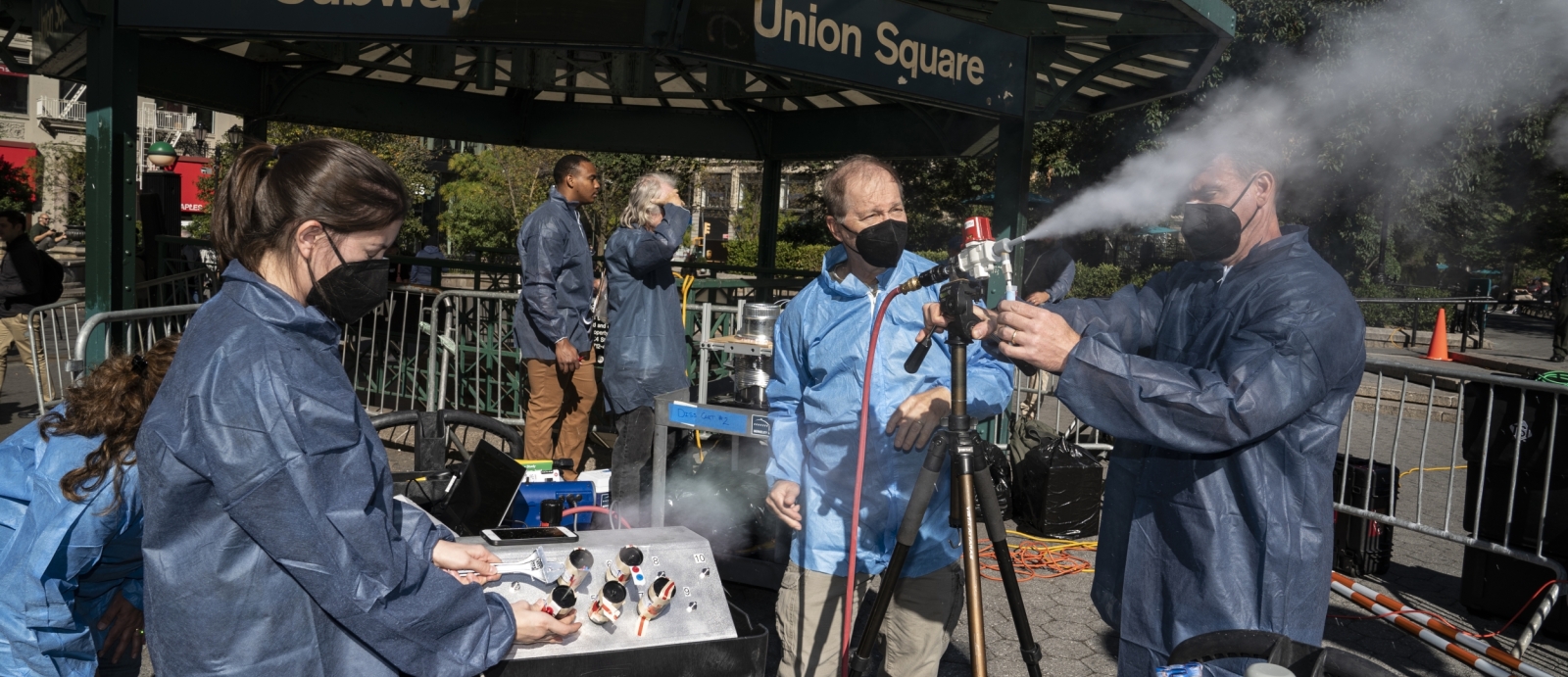 Six scientists in blue lab coats stand in NYC, under a metal gazebo that says "Subway, Union Square" Two of the scientsits operate a small sprayer, on a tripod, that is shooting out a plume of white particles.