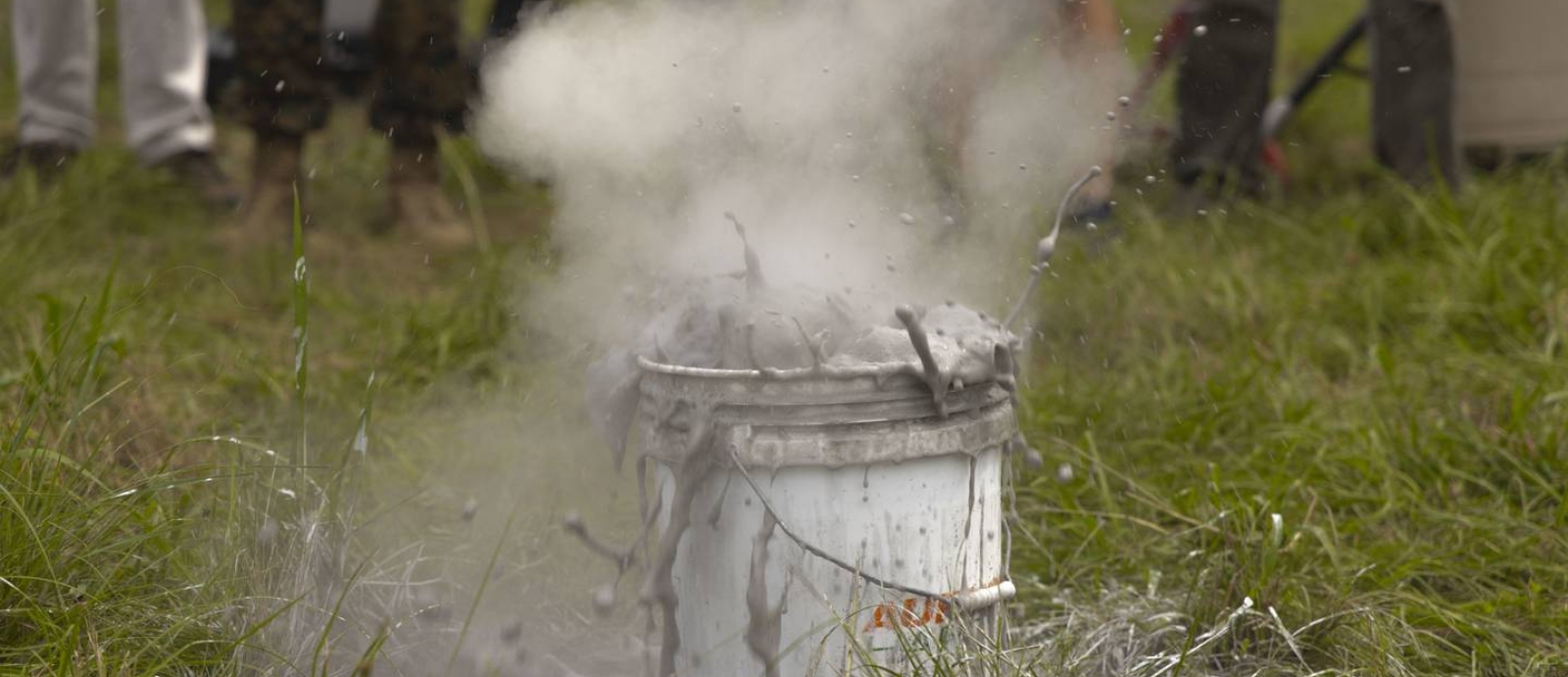 A bucket with silver liquid and gas frothing out of it, on a grassy field.