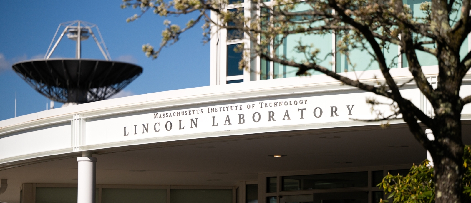 The exterior main entrance of MIT Lincoln Laboratory is shown with a radar antenna in the background.