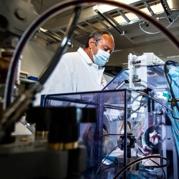 A man stands in a lab wearing a mask, in front of a big box setup where he is testing masks for air filtration. 