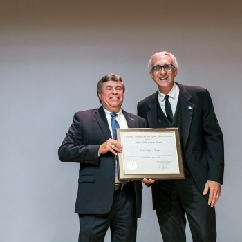 Two men stand next to each other onstage with an award plaque in their hands in front of them. 