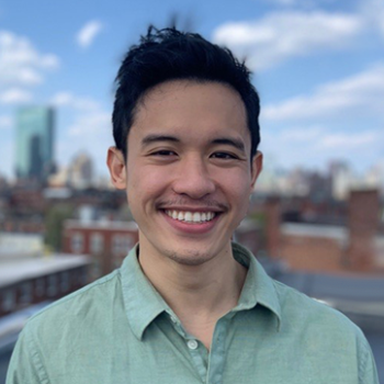 A photo of Mark Hernandez smiling, wearing a green button up shirt. He is outside on a balcony with a view of Boston behind him.