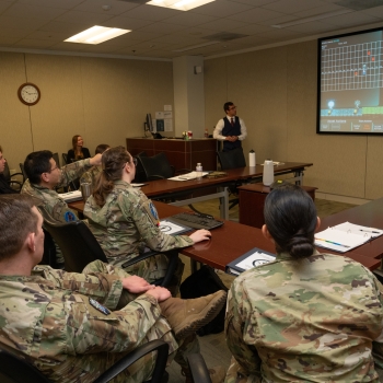 The military interns are gathered in a meeting room and playing the game, which is displayed at the front on a screen.