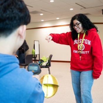 Yelina Britos-Milian is smiling while holding a large gold-colored pendulum. She is wearing a red sweater and another LLRISE student wearing blue looks on in the foreground. 