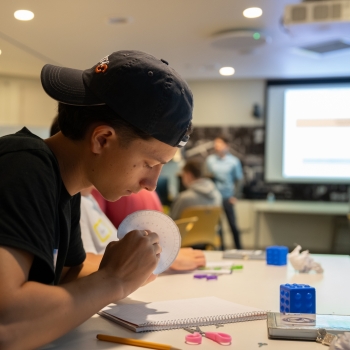 A student sits at a desk in a Beaver Works classroom and is looking at and holding a Caesar Cipher wheel. Other students are sitting in the background doing a similar task. 