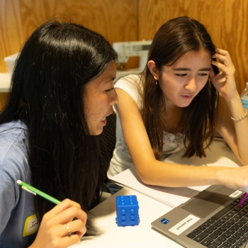 Two students sit together at a classroom desk, looking at a laptop while collaborating on a task