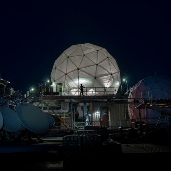 Four satellite dishes and two radomes, each covering satellite communications antennas, are shown on the roof of Lincoln Laboratory among other equipment.