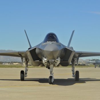 An F-35 aircraft is shown on a runway, with desert mountains in the distance. 