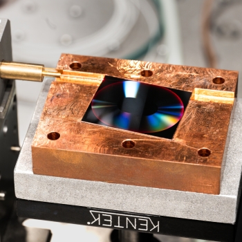 An optical assembly showing a precision mount with brass cylindrical connector attached to a copper block. The block contains a dark optical element with iridescent reflections. 