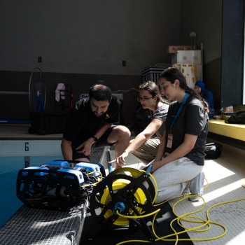 At an indoor pool, two BWSI students confer with a mentor regarding the build of the autonomous underwater vehicle. A third student sits on a bench using her laptop to make programming adjustments for the underwater vehicle.    