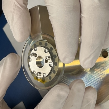 A close-up view of a metallic component of the payload is shown, with a person's gloved hands touching it. 