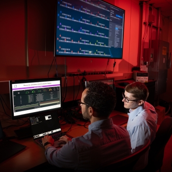 Two researchers sit in front of computer screens in a laboratory, monitoring data representing network performance. 