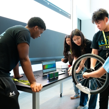 Four students in matching tee shirts from the LLRISE program show a young museum visitor how a self-built radar can detect the movement of a bicycle wheel. 