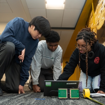 A team of high school students (two boys and one girl) kneel on the floor to test their shoebox-size self-built radar to detect movement down a long hallway.  
