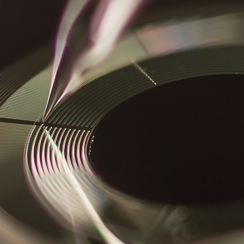 A closely zoomed in photo of a disk with closely space grooves. A sharp point of an instrument is inserted into one of the grooves, where it is dispensing water droplets.