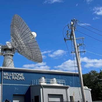 The radar is shown on top of a blue and grey building entitled the Millstone Hill Radar. A bright blue sky is in the radar’s background. 