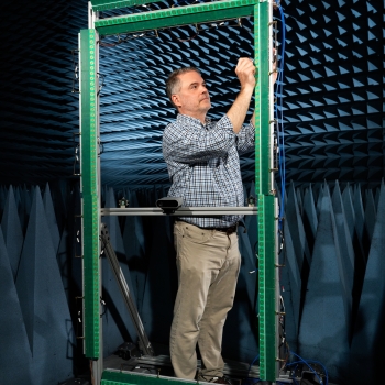 A researcher stands within a rectangular metal frame containing RF antennas and components, surrounding by blue foam within an anechoic “or echo free” chamber. 