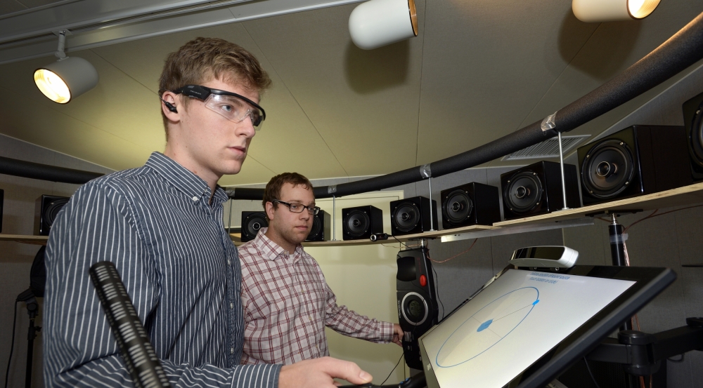 Two staff members stand in a room surrounded by speakers and use a system to select which speaker a sound is coming from.