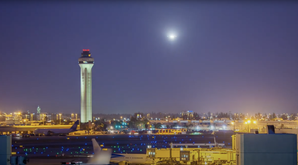 A nighttime photo overlooking an airport runway with several large jets on it, and the illuminated air traffic control tower behind the runway.