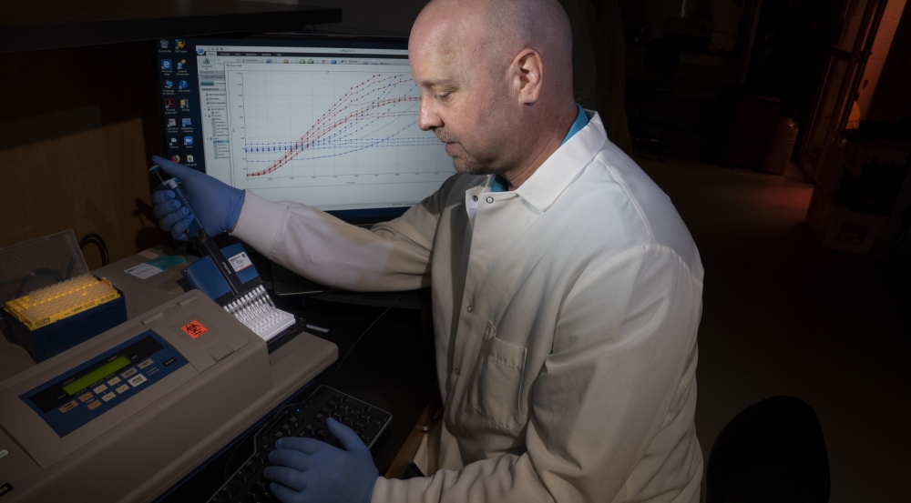 A man in a lab coat puts a vile into a testing machine, with a computer screen showing a graph behind him. 