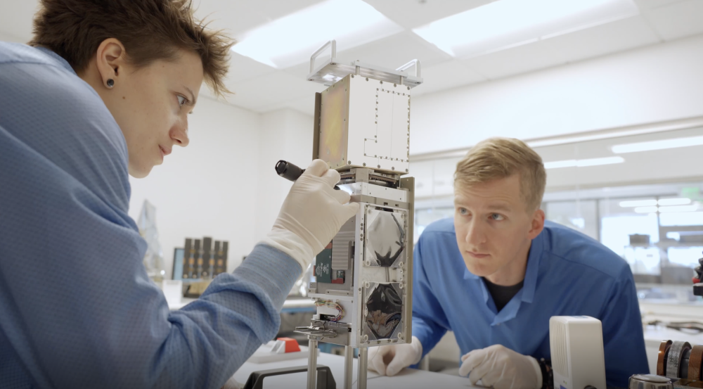 Two researchers inspect a small satellite being assembled on a table. 