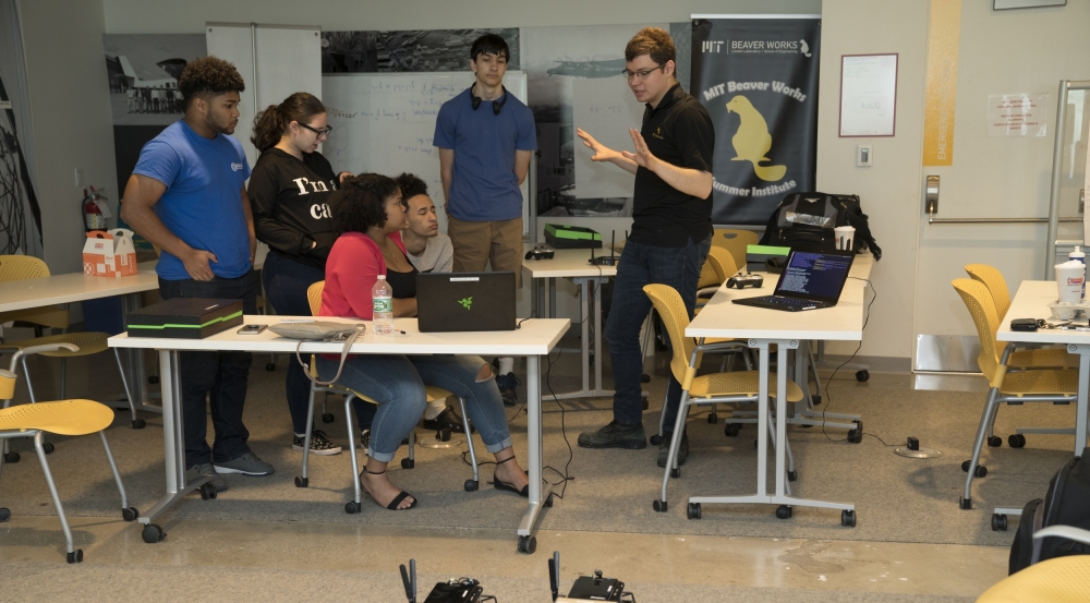 Students sitting around tables discuss the upcoming race surrounded by laptops. Two RC cars are stationed on the floor.