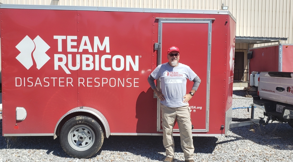 A photo of man in a khakis, a grey tshirt, and red hat standing for a photo in front of a red trailer, with the words Team Rubicon on it. 