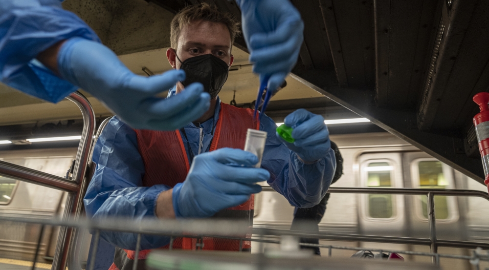 A researcher in an orange vest and blue gloves is putting a small filter into a collection tube (they are helped by their colleague, out of frame except for their hands). They are in a subway station, with a train in view behind them..