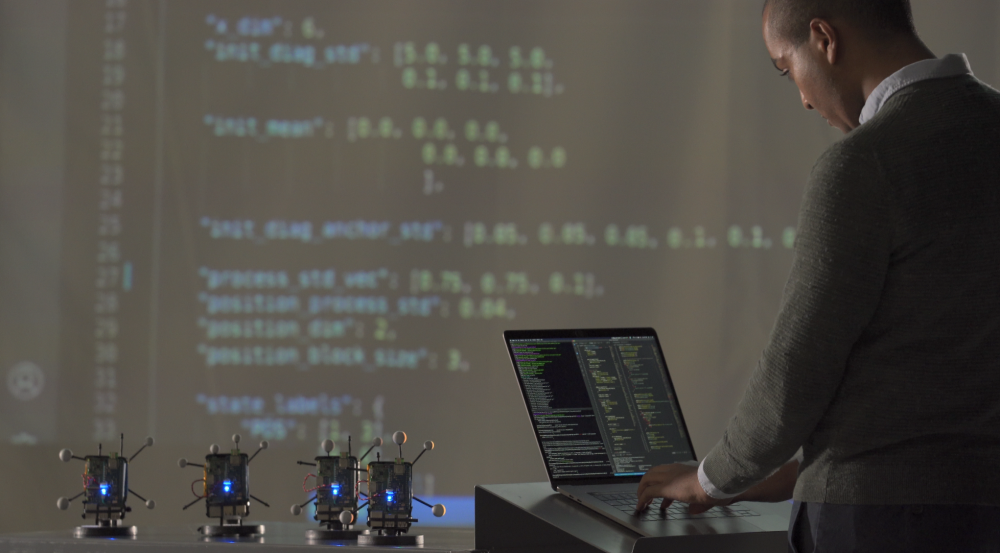 A researcher stands on a laptop. In front of him are a few small radios, and a large projector screen showing data. 