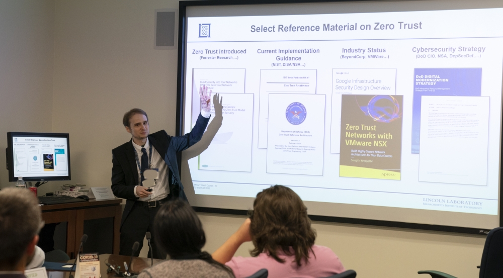 A presenter stands in front of a classroom, pointing to a large screen containing a presentation slide. 