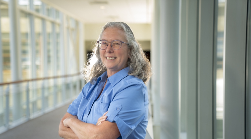 Mary Ellen Zurko poses for a photo in a sunny, glass hallway. She's wearing a light blue shirt and her arms are crossed in front of her.