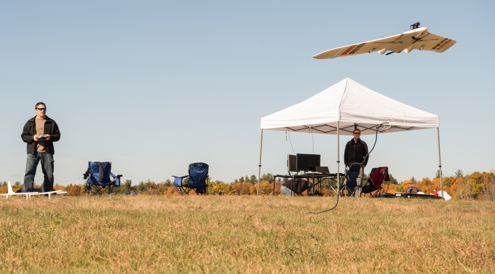 a researcher flies a small drone via remote control. He is standing in field; a tent is set up behind him, where more researchers are working.