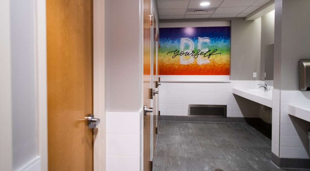 A photo showing the interior of the all-gender bathroom. On the left-hand side are doors to the hard-walled stalls, in the middle is a sign saying "Be Yourself," and to the right are sinks.
