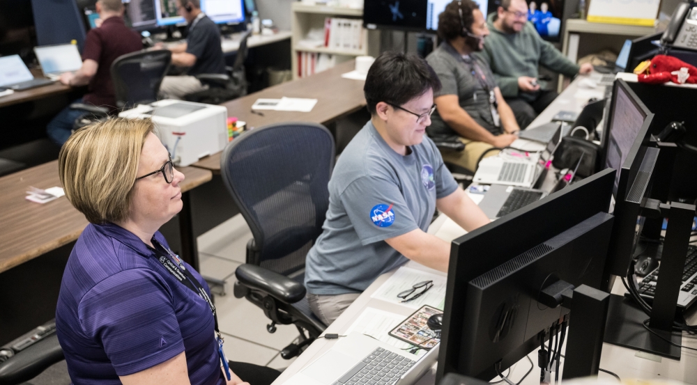 Two people in the foreground and four people in the background sit in front of computer screens.
