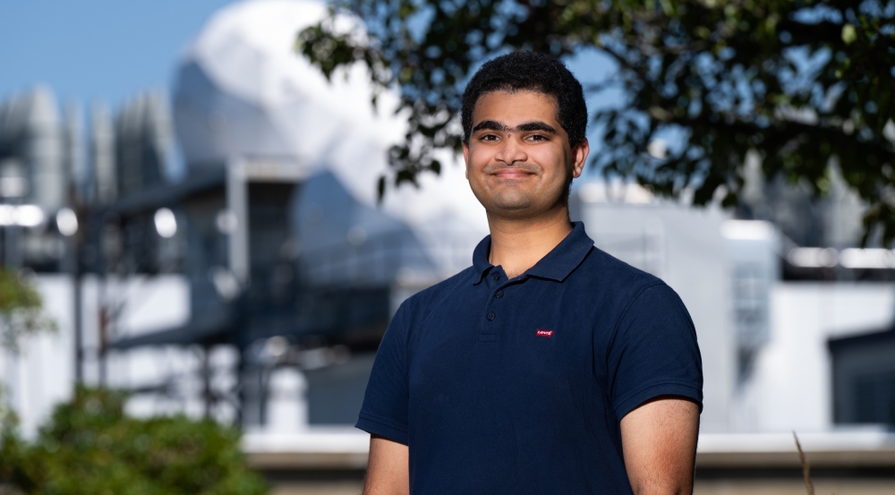 Vivek Jagadeesh smiles for a photo outside of Lincoln Laboratory, with radar antenna radomes in the background. 
