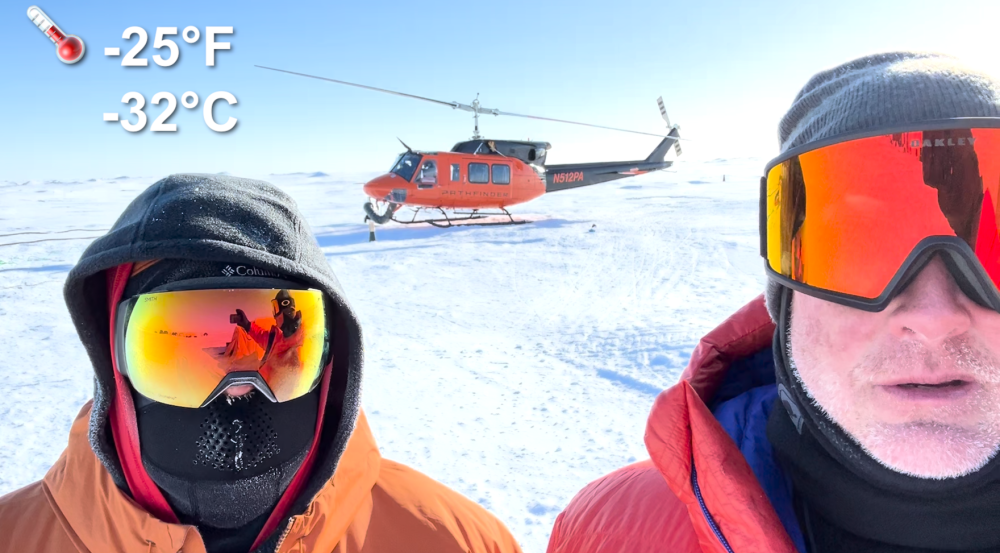 Ben Evans and Dave Whelihan stand bundled up outside in minus 25 degrees Fahrenheit in the Arctic, with a helicopter behind them, as they document their experience in the Arctic.