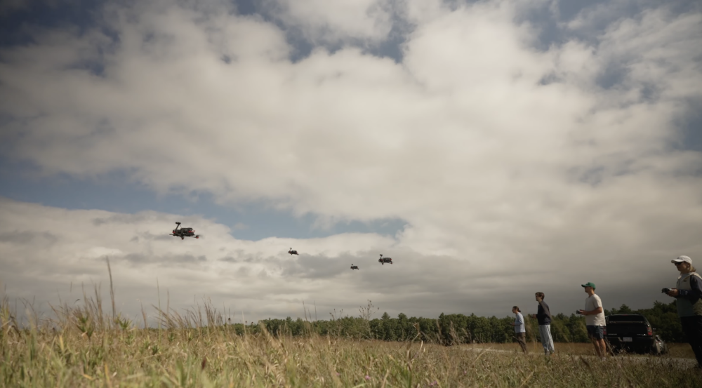 Four staff members stand in a grassy field controlling four drones flying against a cloudy sky.
